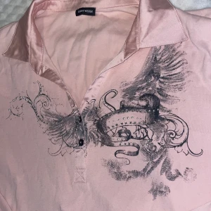 Rosa pikétröja med tryck från Gerry Weber - Snygg ljusrosa pikétröja från Gerry Weber med svart konstnärligt tryck och glittriga detaljer framtill. Tröjan har krage och knappar i satinliknande material samt korta ärmar. Perfekt för dig som gillar unika detaljer och en feminin look.