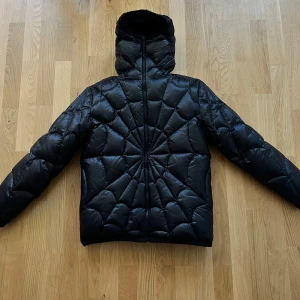 Svart Moncler dunjacka med huva - Säljer en svart dunjacka från Moncler med unik quiltad design som bildar ett spindelmönster . Jackan har huva, hel dragkedja och är lätt glansig. Perfekt för kalla dagar när du vill hålla stilen och värmen. Klassisk Moncler-detalj med serietidningslapp på insidan.