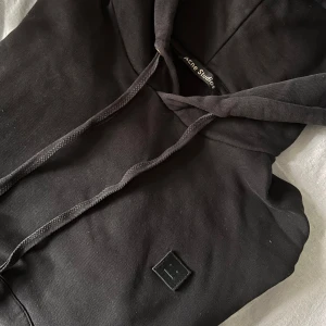 Svart hoodie från Acne Studios - Svart hoodie från Acne Studios med klassisk smiley-patch på bröstet. Tröjan har huva med snörning, magficka och en relaxed passform. 