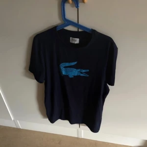  Lacoste Sport t-shirt med blå logga - Snygg svart t-shirt från Lacoste Sport med klassisk blå krokodil-logga på bröstet. T-shirten är kortärmad och har en rund halsringning. Perfekt för dig som gillar stilrena plagg med sportig touch.