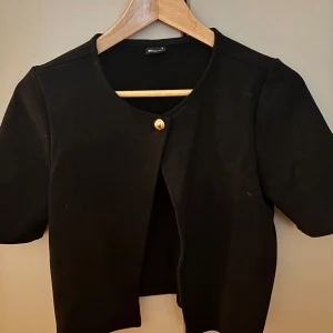 Svart kort cardigan från Gina Tricot - Kort svart cardigan jacka från Gina Tricot med rund halsringning och en snygg guldig knapp framtill. Helt ny och aldrig använd. Superfin över ett linne tillexempel. Väldigt trendig.