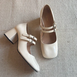 Krämvita pumps från & Other Stories - Snygga pumps i krämvitt lack från & Other Stories med fyrkantig tå, dubbla remmar med silverspännen och stabil blockklack. Skorna har en retrovibe och passar perfekt till både jeans och klänning. Materialet är glansigt syntet som ger en lyxig känsla.