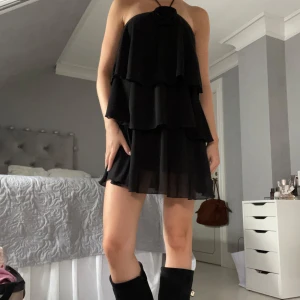 Svart volangklänning med halterneck - Supersöt svart kortklänning med flera volanger och halterneck med en ros längst upp. Prislapp på 