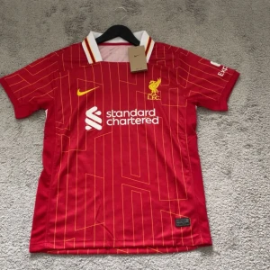 Liverpool FC röd matchtröja Nike - Liverpool FCs officiella matchtröja från Nike i rött med gula och vita detaljer. Tröjan har tunna gula linjer i ett geometriskt mönster, vit krage med röda och gula ränder samt klubbmärke och sponsortryck på bröstet. Tillverkad i lätt och ventilerande material.