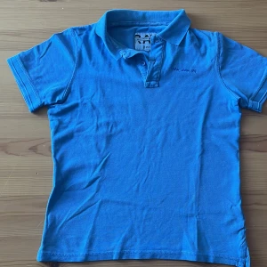 Blå pikétröja från RAW by FILA - Snygg blå pikétröja från RAW by FILA med klassisk krage och korta ärmar. Tröjan har broderad text på bröstet och knäppning vid halsen. Perfekt för en chill och sportig look. Tillverkad i mjuk bomull som känns skön mot huden.