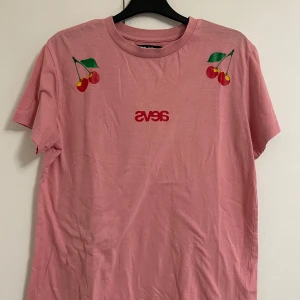 Rosa t-shirt med körsbär från Svea - Säljer en rosa t-shirt från Svea med tryckta körsbär på axlarna och röd text på bröstet. T-shirten är i mjuk bomull och har klassisk passform med rund hals och korta ärmar. 