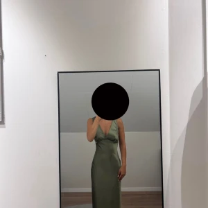 Olivgrön långklänning med v-ringning - Snygg olivgrön långklänning i satinkänsla med djup v-ringning och ärmlös design. Klänningen har en figurnära passform upptill och faller snyggt längs kroppen. Perfekt för dig som vill ha en elegant och stilren look.