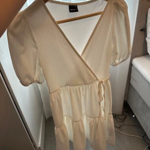 Vit omlottklänning från Gina Tricot - Superfin vit omlottklänning från Gina Tricot med puffärmar och knyt i midjan. Klänningen har v-ringning, volangdetaljer och är tillverkad i ett lätt, strukturerat tyg. Perfekt för sommarens alla tillfällen och ger en fräsch och luftig känsla.