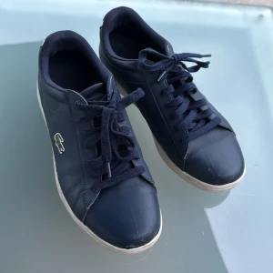 Mörkblå sneakers från Lacoste - Snygga mörkblå sneakers från Lacoste med klassisk krokodillogga på sidan och vit detalj på hälen. Skorna har snörning, platt sula och är tillverkade i skinn med textilfoder. Perfekta för en clean och sportig stil.