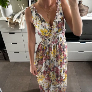 Blommig långklänning från Redoute - Superfin långklänning med blommigt mönster i gult, rosa och lila på vit botten. Djup v-ringning, smock i midjan och volangkant vid axlarna. Perfekt för sommarens alla tillfällen. Lätt och luftig känsla.