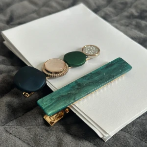 Set med två trendiga hårspännen - Säljer två snygga hårspännen – ett med fyra runda dekorationer i mörkblått, grönt, beige och glittrigt silver, infattade i guldfärgade detaljer, samt ett rektangulärt spänne i marmorerad grön nyans med guldklämma. Perfekt för att lyfta din look.