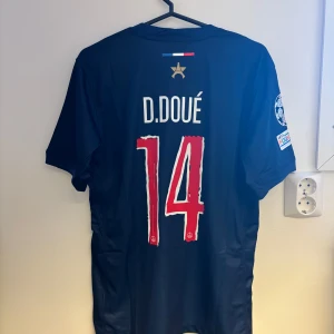 PSG matchtröja D. Doué #14 Nike S - Paris Saint-Germain fotbollströja med D. Doué och nummer 14 på ryggen. Mörkblå bas med bred röd och vit rand framtill, PSG-emblem och Nike-logga. Qatar Airways sponsortryck och Champions League-märke på ärmen. Tillverkad i lätt och ventilerande material.