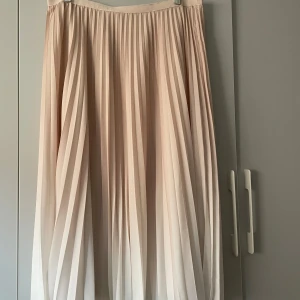 Plisserad beige långkjol French Connection - Superfin plisserad långkjol från French Connection i en ljus beige ton. Kjolen har ett flowigt fall och resår i midjan för extra komfort. Perfekt att styla med sneakers eller boots för en chill och trendig look.