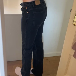 Svarta Weekday Pin Mid Straight jeans - Säljer ett par svarta jeans från Weekday, modell Pin Mid Straight. Klassisk femficksdesign med raka ben och midja som sitter mitt på höften. Jeansen är i bomull och har snygga kopparfärgade nitar. Perfekta till en clean och stilren look. Storlek W26 L33