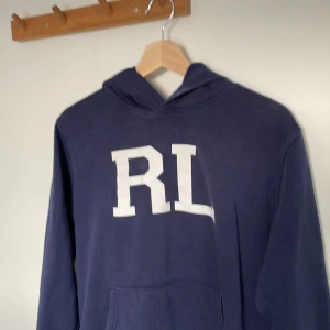 Ralph lauren hoodie - Säljer en mörkblå hoodie från Ralph Lauren med stora vita RL-bokstäver på bröstet. Tröjan har huva och känguruficka framtill, perfekt för en chill och sportig look. Materialet är mjuk bomull och passformen är normal. Fråga om det är något du undrar om.
