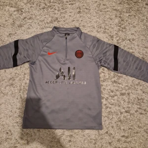 Grå PSG träningströja från Nike - Snygg grå långärmad träningströja från Nike med PSG-logga och röd swoosh på bröstet. Tröjan har svart detalj på ärmarna, halv dragkedja och tryckt sponsorlogga framtill. Perfekt för fotbollsträning eller chill.