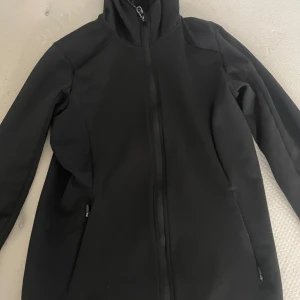 Svart zip up träningströja med huva - Svart träningströja med hel dragkedja och huva. Jackan har en clean look, långa ärmar och två fickor med dragkedja. Perfekt för dig som vill ha en enkel och stilren jacka till vardags. Materialet känns lätt och smidigt.