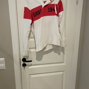 Vit och röd hoodie från GANT XS - Snygg hoodie från GANT i vitt med breda röda ränder och tryck med texten 'GANT 1949' fram och 'NEW HAVEN' bak. Tröjan har huva med snörning, långa ärmar och är gjord i mjuk bomull. Perfekt för en sportig och avslappnad look. (Om du vill köpa köp på Vinted brage2011)