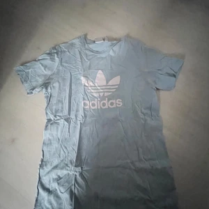 Ljusblå Adidas t-shirt med logga - Snygg ljusblå t-shirt från Adidas med den klassiska vita treklöversloggan tryckt på bröstet. T-shirten är kortärmad och har en avslappnad passform. Perfekt för dig som gillar sportiga och stilrena plagg.