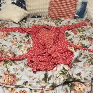 Röd blommig omlottklänning Amelie & Me - Superfin röd omlottklänning från Amelie & Me med vitt blommönster. Klänningen har volangdetaljer, långa ärmar och knytband i midjan. Perfekt för dig som vill ha en feminin och trendig look med extra flow.