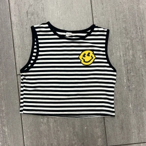 Randig croppad linnetopp från SHEIN - Svartvit randig croppad linnetopp från SHEIN med ett stort gult smileytryck på bröstet. Toppen är ärmlös och har ribbad struktur, perfekt för en chill och lekfull stil. Passar dig som gillar att sticka ut med coola detaljer.
