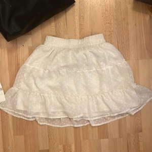 Vit volangkjol med prickar - Supersöt vit kortkjol med volanger och diskret prickigt mönster. Kjolen har resår i midjan för skön passform och är gjord i ett lätt, luftigt material som passar perfekt till varmare dagar. Den har en flowy känsla som ger en gullig och trendig look.