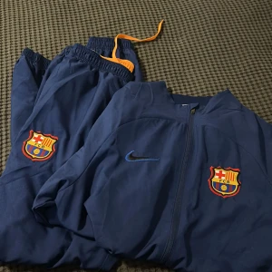 Blå Nike FC Barcelona träningsoverall - Säljer en blå teäbubgsoverall från Nike med FC Barcelona-logga på bröstet och dragkedja framtill. Jackan har lång ärm och är tillverkad i lätt polyester, perfekt för fotbollsträning eller när du vill representera Barca. Snygg broderad Nike-swoosh på ena sidan. Byxorna har även de snygg broderad logga. 