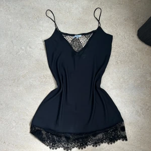 Svart linnetopp med spetsdetaljer - Supersnygg svart linnetopp från Vila med tunna axelband och v-ringning. Toppen har fina spetsdetaljer både vid urringningen och längs nederkanten som ger en extra touch. Perfekt att styla lager på lager eller som den är för en chic look.