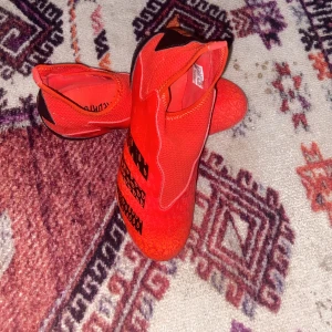 Röda Adidas Predator fotbollsskor - Snygga röda Adidas Predator fotbollsskor med svarta detaljer och mönster på ovandelen. Skorna har en strumpliknande passform utan snörning och är tillverkade i syntetmaterial för bästa grepp och känsla på planen.