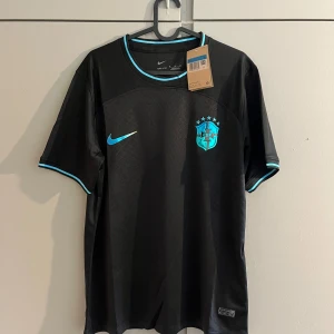 Svart Brasilien fotbollströja Nike - Snygg svart Brasilien fotbollströja från Nike med blå detaljer och CBF-emblem på bröstet. Tröjan har korta ärmar, rund hals och är tillverkad i ett lätt, andningsbart material. Perfekt för dig som vill sticka ut på planen eller på läktaren.