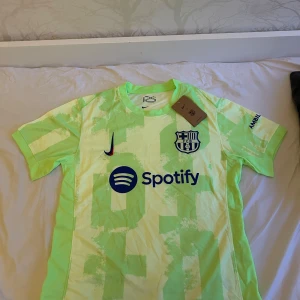 Barcelona Nike Dri-FIT fotbollströja S - Barcelona fotbollströja från Nike i Dri-FIT-material. Tröjan är ljusgrön med gula inslag och har ett unikt mönster. Klubblogga, Nike-logga och Spotify-tryck på bröstet, samt UNHCR-tryck på ryggen. Kortärmad och perfekt för dig som älskar Barca.