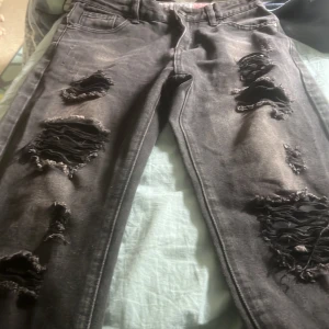 Svarta ripped jeans med dragkedja - Säljer ett par svarta jeans med rejält slitna och trasiga detaljer på båda benen. Jeansen har dragkedja och knapp framtill, klassisk femficksmodell och coolt, edgy vibe. Perfekta för dig som gillar streetstyle och vill sticka ut.