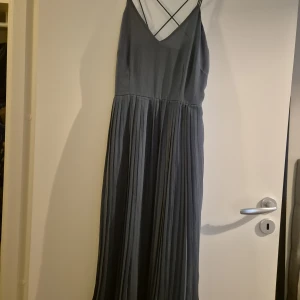 Grå långklänning från NA-KD - Supersnygg grå långklänning från NA-KD med tunna axelband som korsas i ryggen och v-ringning. Kjolen är plisserad för en extra flowy känsla. Perfekt för dig som gillar stilrena och eleganta vibes.