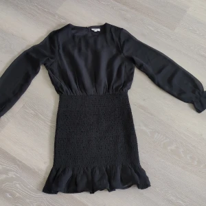 Svart kort klänning från KappAhl - Supersnygg svart kort klänning från KappAhl med långa, lätt transparenta ärmar och smockad midja. Kjolen har volangkant nertill och toppen är i ett mjukt, flowy tyg. Perfekt för dig som gillar stilrena och trendiga plagg.