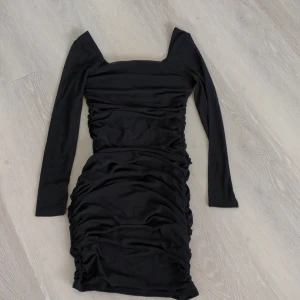 Svart tight kortklänning med meshärmar - Snygg svart kortklänning med långa meshärmar och rynkade detaljer längs sidorna. Klänningen har en fyrkantig ringning och är figurnära, perfekt för dig som gillar en trendig och stilren look. Tillverkad i stretchigt material med polyamid.