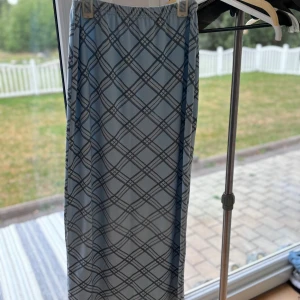 Ljusblå långkjol med rutigt mönster - Långkjol i ljusblått med svart rutigt mönster. Kjolen har resår i midjan för en bekväm passform och är rak i modellen. Perfekt för dig som gillar en stilren och trendig look.