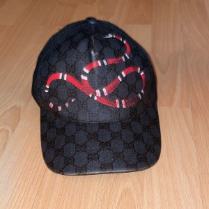 keps med orm och GG-mönster - Svart keps från med klassiskt GG-monogram över hela kepsen och en röd orm tryckt framtill. Baksidan har mesh-tyg och justerbart band med röd och blå rand. Snygg och unik accessoar för dig som vill sticka ut.