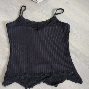 Svart ribbad linnetopp med spets -  svart ribbad linnetopp från Romwe med tunna axelband och spetsdetaljer både upptill och nertill. Toppen har en smickrande v-ringning och är perfekt för att skapa en cool lager-på-lager-look eller bära som den är.