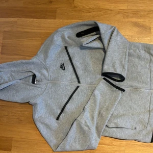 Grå Nike hoodiejacka XS - Snygg grå hoodiejacka från Nike i storlek XS Jackan har hel dragkedja, svarta detaljer och fickor med dragkedja. Perfekt för en sportig look och chill vardagsstil. Mjuk och skön med huva och lång ärm.