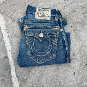 True Religion Jeans - Säljer ett par blå jeans från True Religion med ikoniska kontrastsömmar och stora bakfickor med snygga detaljer. Jeansen har klassisk passform, slitningar och tydliga logoknappar. Perfekta för dig som gillar streetwear och vill sticka ut. (Extremt slitna)