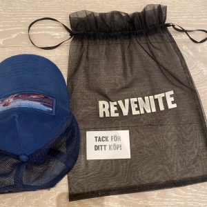 Blå keps från Revenite med motiv - Snygg blå keps från Revenite med mesh på sidorna och justerbar snapback. Framtill finns ett tryckt motiv med bil och kvinna vid hamn. Kepsen har broderad logga på insidan och levereras med originalpåse.