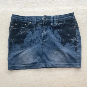 Jeanskjol med svarta paljetter - Snygg jeanskjol i blå denim med svarta paljettdetaljer upptill och på fickorna. Kjolen har klassisk femficksdesign, bälteshällor och dragkedja fram. Perfekt för dig som vill sticka ut med lite extra bling i vardagen.