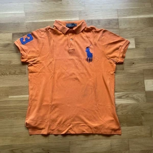 Ralph Lauren piké  - Orange pikétröja från Polo Ralph Lauren med blå broderad logga på bröstet och blå siffra på ärmen. Klassisk krage och knappar framtill. Tillverkad i bomull och har korta ärmar. Perfekt för en chill och sportig look. I använt skick med lite blekt färg och ett litet hål som syns på sista bilden :)