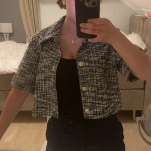 Rutig croppad overshirt från H&M - Snygg croppad overshirt från H&M i svartvitt rutigt mönster. Jackan har korta ärmar, klassisk krage och guldiga knappar framtill. Perfekt för att lyfta din outfit med en trendig touch. Aldrig använd !