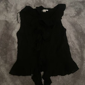 Svart ärmlös blus med volang - Säljer en svart ärmlös blus med volangdetaljer längs kragen och framtill. Blusen har en lös passform och knytband framtill som ger en extra touch. Storlek 158-164