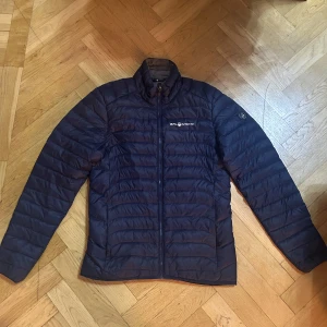 Mörkblå Sail Racing dunjacka - Snygg mörkblå dunjacka från Sail Racing med quiltad design och dragkedja framtill. Jackan har hög krage, lång ärm och logga på bröstet samt ärmen. Perfekt för kyliga dagar och enkel att matcha med det mesta.