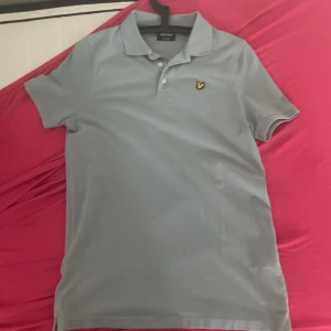 Ljusgrå pikétröja från Lyle & Scott - Snygg ljusgrå pikétröja från Lyle & Scott med klassisk krage, korta ärmar och knappar framtill. Tröjan har det ikoniska gula örnloggan broderad på bröstet. Perfekt för en clean och stilren look.