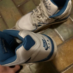 Nike Air Flight vita och blå sneakers - Snygga Nike Air Flight sneakers i vitt och blått med klassisk retrostil. Skorna har snörning, rund tå och är tillverkade i skinn och syntet. Perfekta för dig som gillar old school vibes och vill sticka ut med en ikonisk modell.