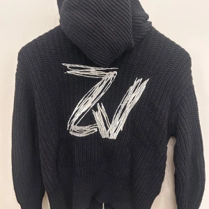 Svart stickad hoodie från Zadig & Voltaire - Svart chunky stickad hoodie från Zadig & Voltaire med dragkedja framtill och stor broderad ZV-logga i vitt på ryggen. Tröjan har huva, långa ärmar och en cool oversized känsla. 