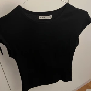 Svart topp från Pull&Bear - En svart, enkel topp från Pull&Bear. Modellen har korta ärmar och rund halsringning. Perfekt att matcha med höga jeans eller kjol för en clean look. Tunn och mjuk i materialet, passar året runt. Storlek M